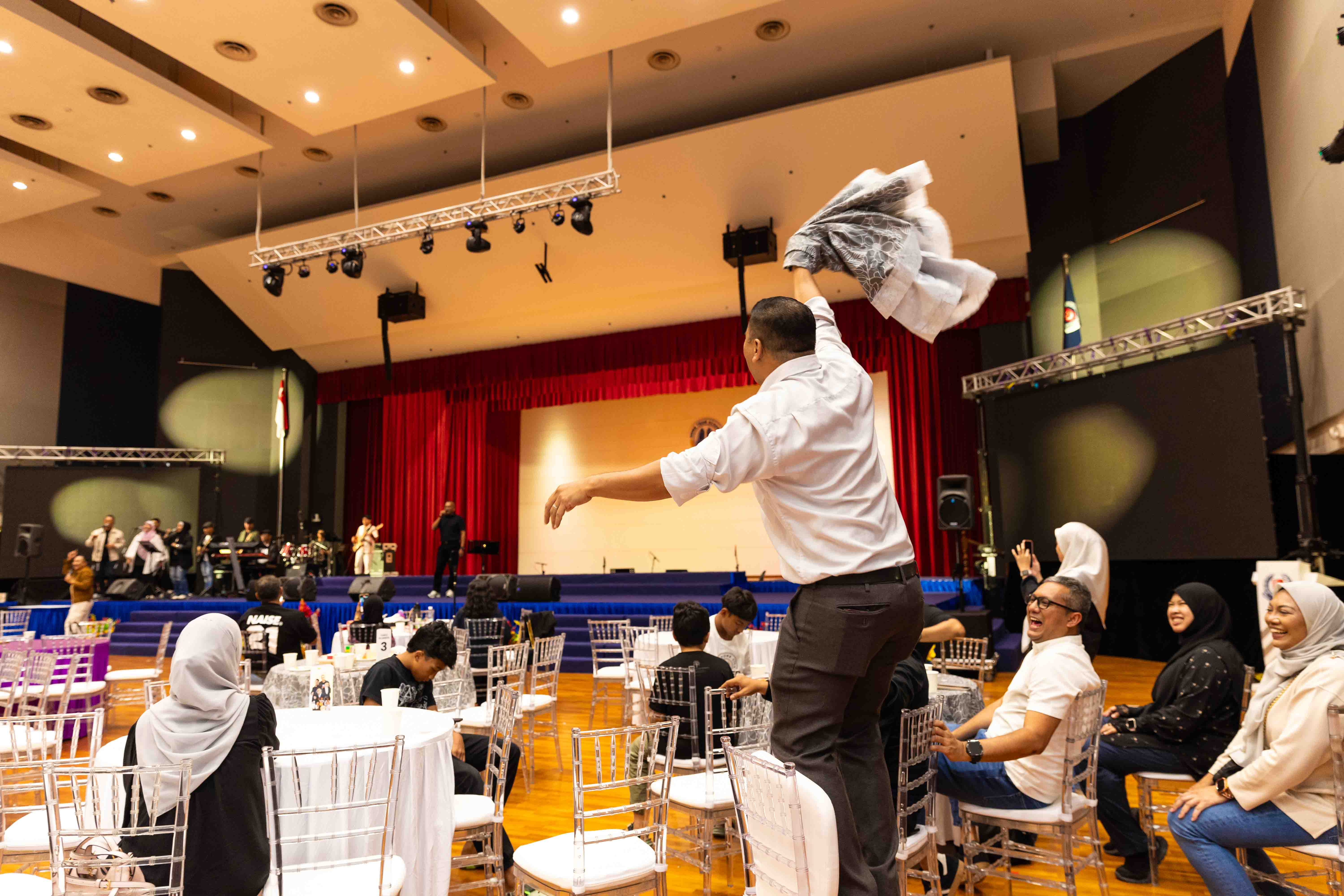 An attendee stands on a chair waving a jacket while others laugh and cheer during the performance.